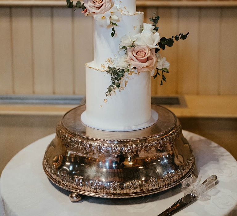 Three tier tall white iced wedding cake with gold flake decorations and pink and white roses with pampas grass and foliage on a tree trunk cake stand 