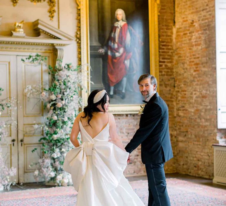 Bride in sleeveless satin wedding dress with large accent bow on the back walking with groom in classic black tuxedo, black bowtie and boutonniere    