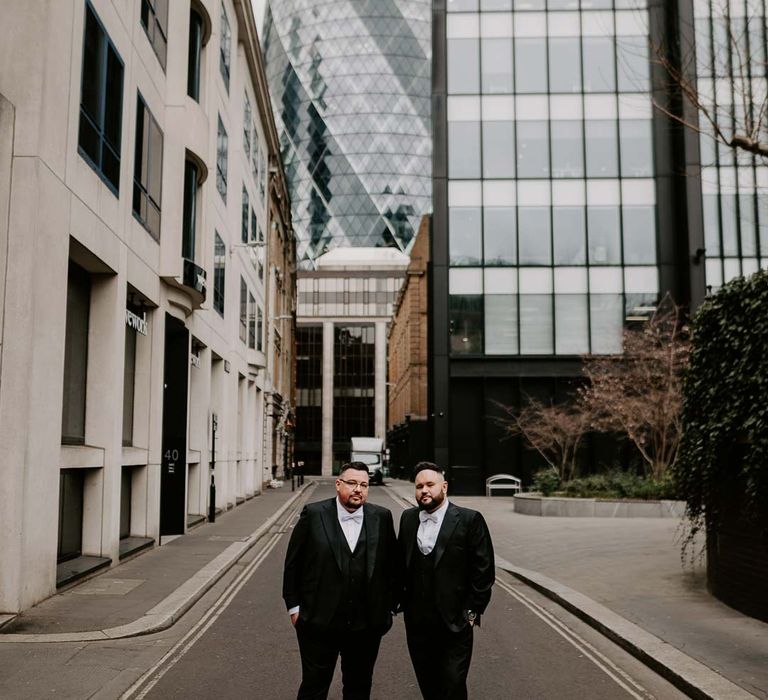 Grooms in matching three piece black tuxedos with off white bow ties, white garden rose, lavender twig and dried foliage boutonnieres and grooms Louboutins shoes standing outside of The Gherkin wedding venue in London 