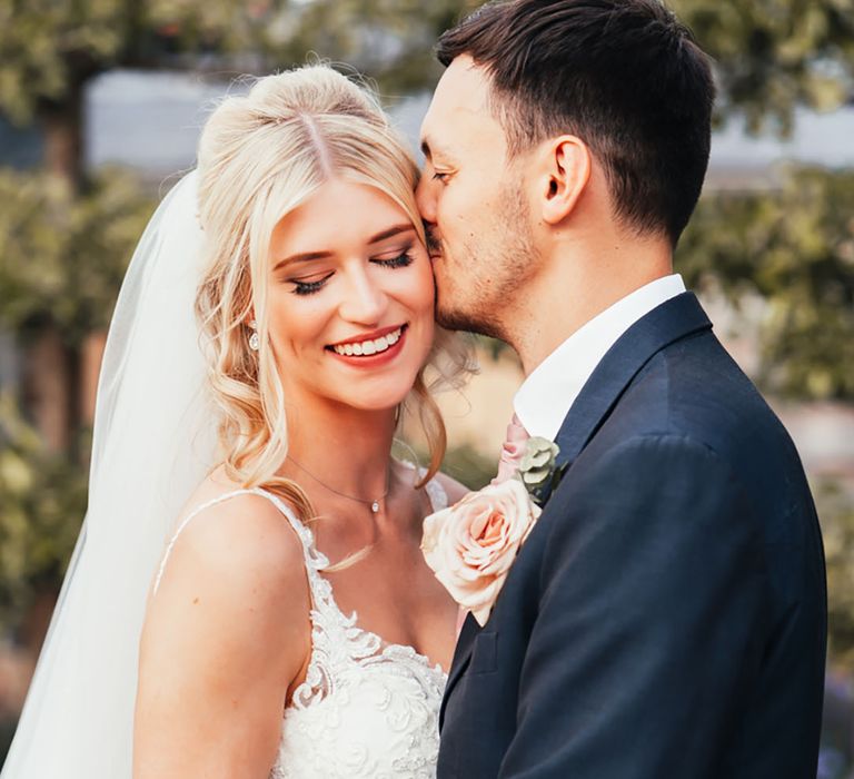 The groom in a navy suit kissing the bride on the forehead who wears a lace wedding dress and veil 