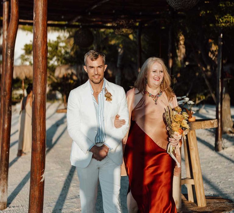 Groom walking down the aisle wearing white mens wedding suit, light blue and white striped shirt, dusky orange garden rose and dried flower boutonniere, white suit trousers and gold jewellery with wedding guest in split style burgundy and dusky orange sleeveless dress 