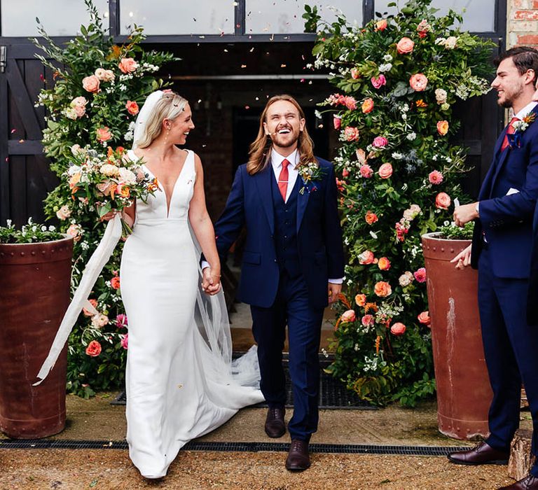 Groom in three piece blue suit walks out of the ceremony to confetti with the bride in a Savin London wedding dress 