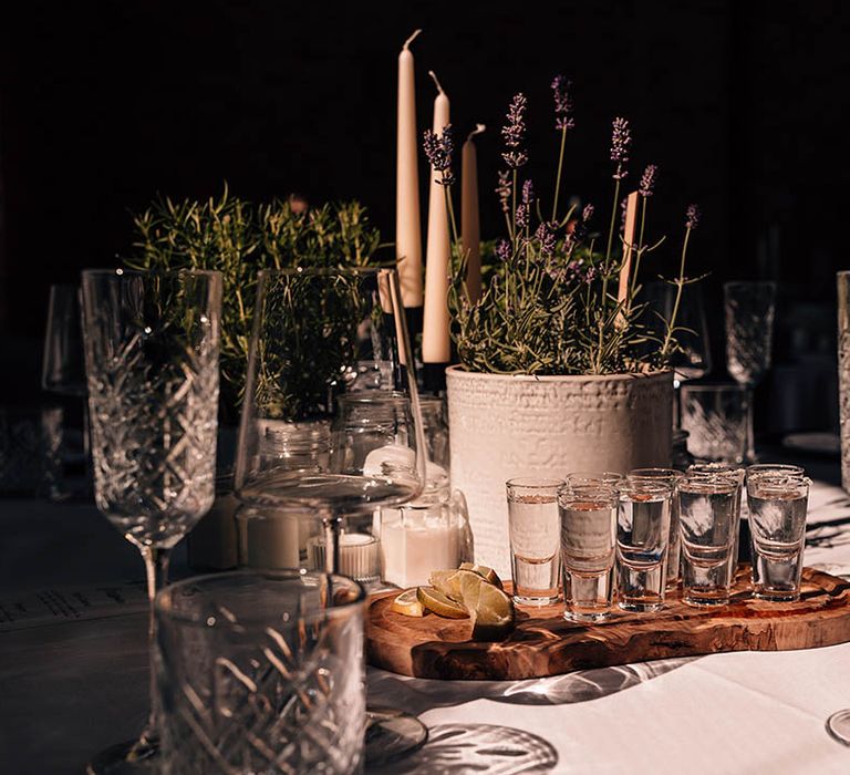 A wooden tray of shots with slices of fruit for the wedding guests on the tables with potted plants of lavender and rosemary with candles 