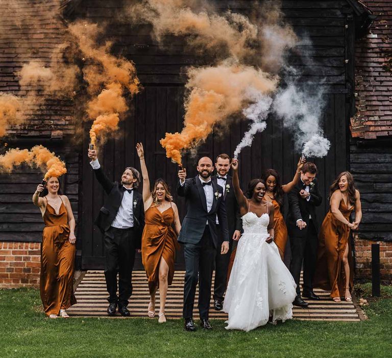 Bride in sweetheart neckline lace wedding dress with detachable off the shoulder sleeves, groom in classic black three piece grooms suit with bow tie and white rose and foliage boutonniere in church, bridesmaids wearing strappy sleeveless front slit burnt orange bridesmaid dresses holding foliage bouquets and groomsmen wearing classic black suits doing burnt orange and white smoke flares at Lains Barn wedding