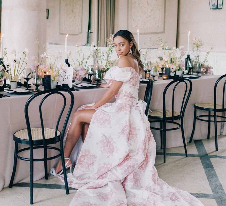 Bride in off the shoulder white and pink floral patterned bridal gown with black bow and front slit sitting at Notan wedding tablescape consisting of light pink wedding tablecloth, white poppies, light pink Persian buttercups, baby's-breath and foliage centrepieces, black artistic candlestick holders, white tapered candles, black crockery and monochrome wedding stationery at Settrington Orangery