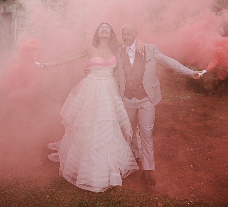 bride in a strapless wedding dress and groom in a grey suit waving pink smoke bombs at italian villa wedding