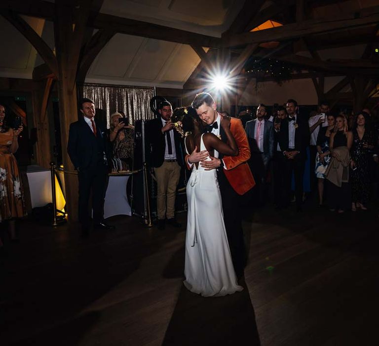 Bride in low back satin look wedding dress with 3D floral applique train slow dancing with groom in crushed burnt orange velvet grooms blazer, black bowtie and black suit trousers to their first dance wedding song