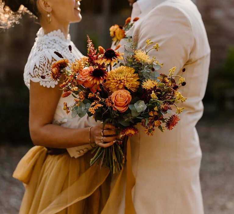 Groom in cream morning suit embracing bride in white mesh lace bridal crop top and yellow wedding skirt holding dried autumnal wedding bouquet with sunflowers, garden roses, pampas grass, eucalyptus, yellow tall kangaroo paws, orange osmanthus flowers, foliage and dried flowers at Broadfield Court wedding