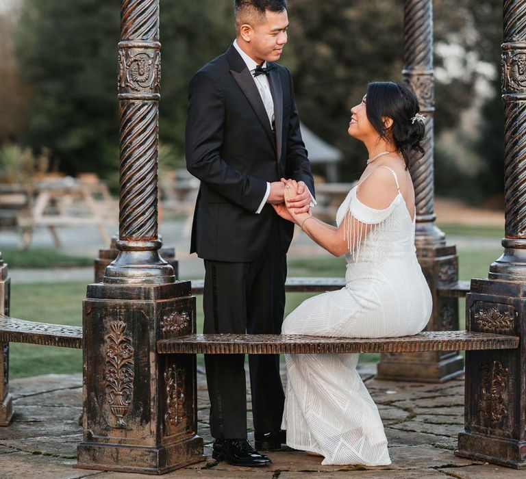 Bride in white beaded and sequined slip wedding dress and groom in classic tux embracing under pergola at Bron Eifion wedding venue for Burmese wedding