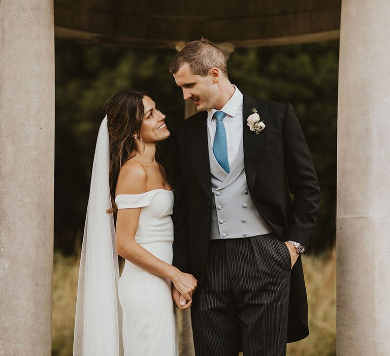 Groom in light grey waistcoat and light blue tie with black morning suit with the bride in an elegant off the shoulder gown 