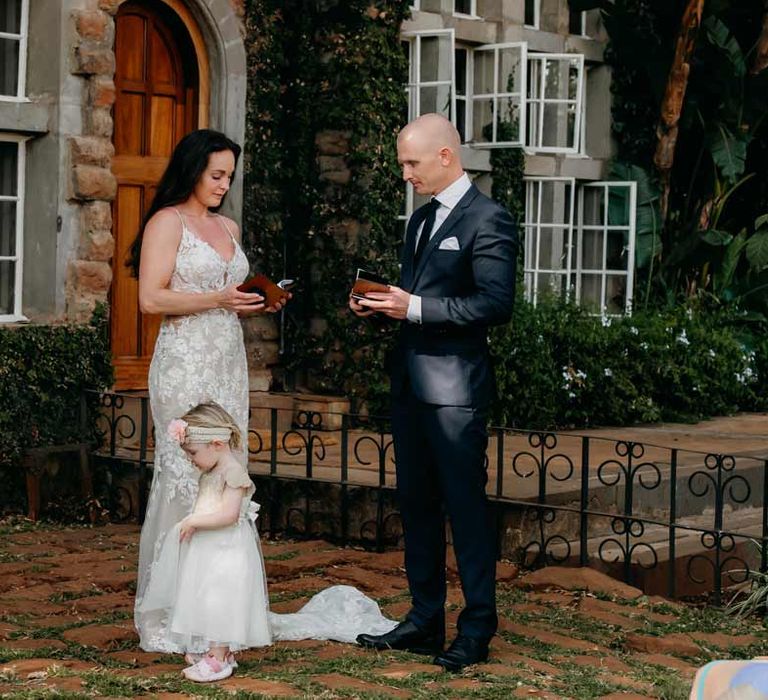 Bride in lace sleeveless wedding dress with puddle train standing with groom in classic black grooms suit and child in off-white dress and sequin headband with pink carnation reading vows