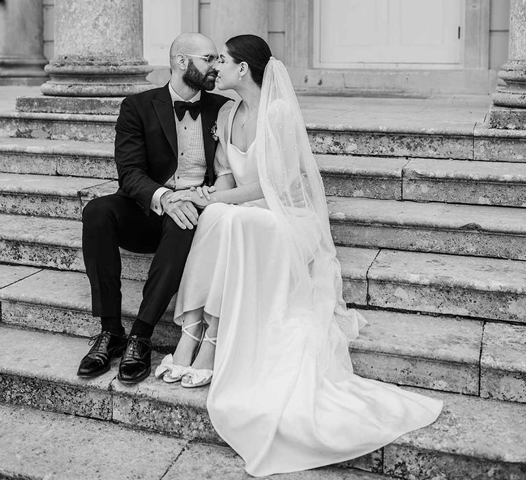 Bride in short sheer puff sleeve wedding dress with square neck and wrist loop with sheer pearl embellished bridal veil kissing groom in classic black tuxedo with bowtie and wildflower boutonniere sitting on the steps of Buxted Park