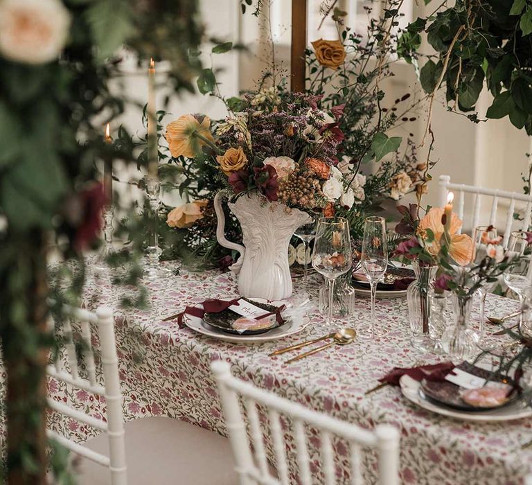 Botanical wedding tablescape with patterned floral tablecloth, twisted tapered candles, art deco vases with dried wildflower wedding centrepieces surrounded by suspended wildflower and foliage arrangements in large gold vases at Buxted Park