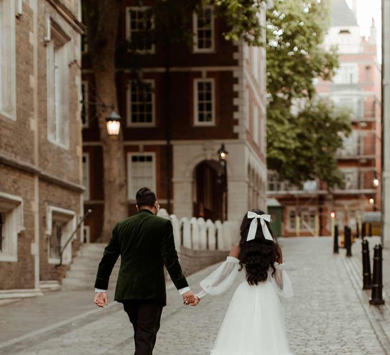 Groom in velvet suit and bride in off the shoulder layered tulle wedding dress and large wedding bow hair accessory walking through the streets of London