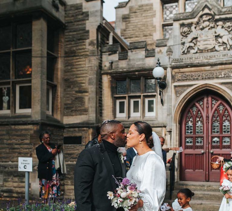 Bride carrying a pink bouquet in a long balloon sleeve satin wedding dress sharing a kiss with the groom in black suit with their kids running around 