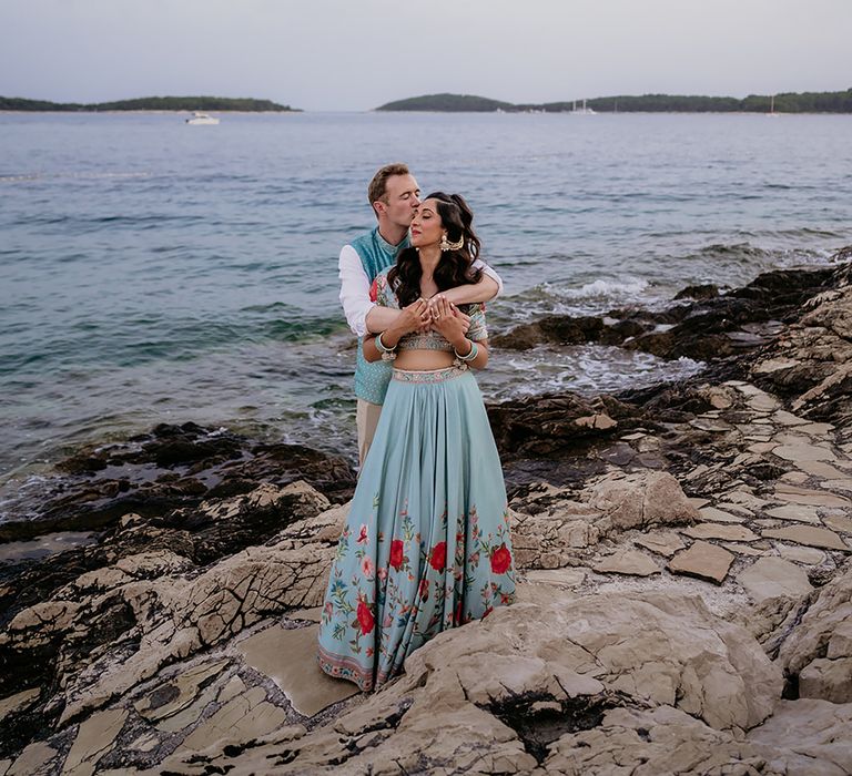 Groom embracing bride in traditional turqouise and pink floral South Asian wedding outfits on rocks near ocean in Hvar, Croatia