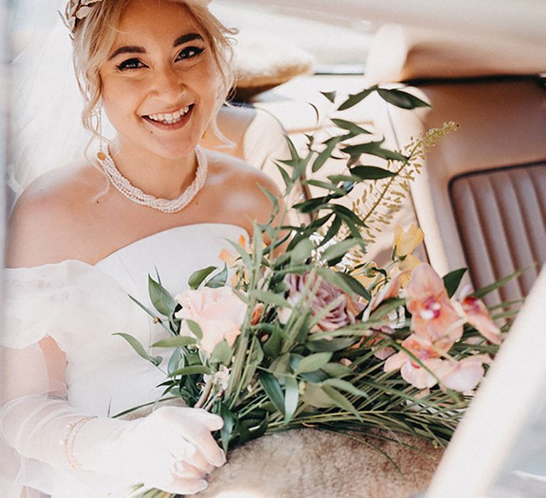 Bride wearing statement necklace to complement her off the shoulder gown sitting in the wedding car 