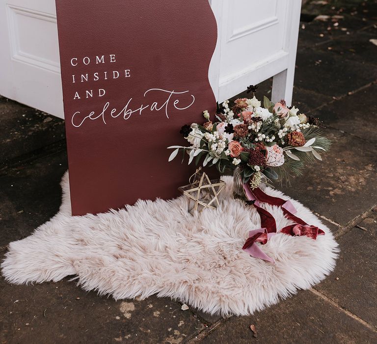 Burgundy wedding welcome sign on furry rug with the bride's winter wedding bouquet with red and pink flowers tied with velvet ribbon