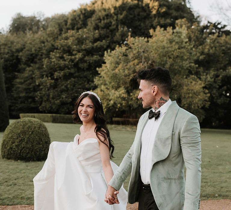 Bride wearing gold bow wedding shoes in Pronovias gown with pearl jewellery walking with groom in three piece green suit with bow tie 