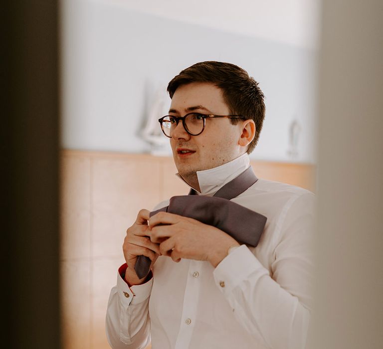 Groom wearing a white shirt puts on his purple tie as he gets ready for the wedding day 