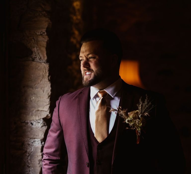 Smiling groom in burgundy Marc Darcy suit looks out the window