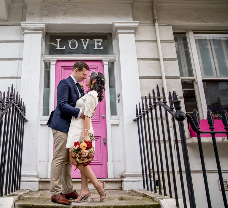 Bride in a short registry wedding dress kissing her groom in front of a pink door with LOVE sign 