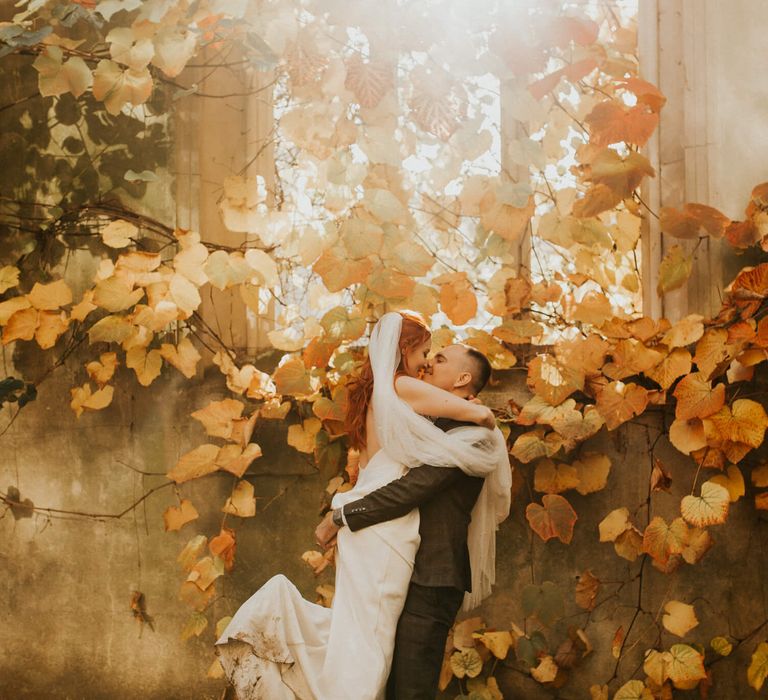 Groom holding his bride surrounded by autumnal leaves bride wears simple white wedding gown and converse 