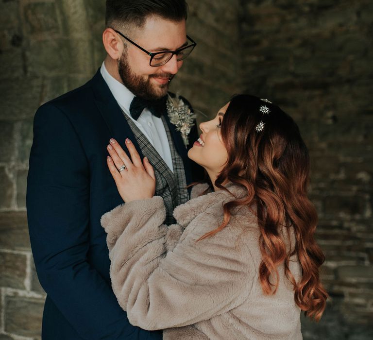 Bride in fur coat and celestial headband smiles up at groom in navy suit and tartan waistcoat