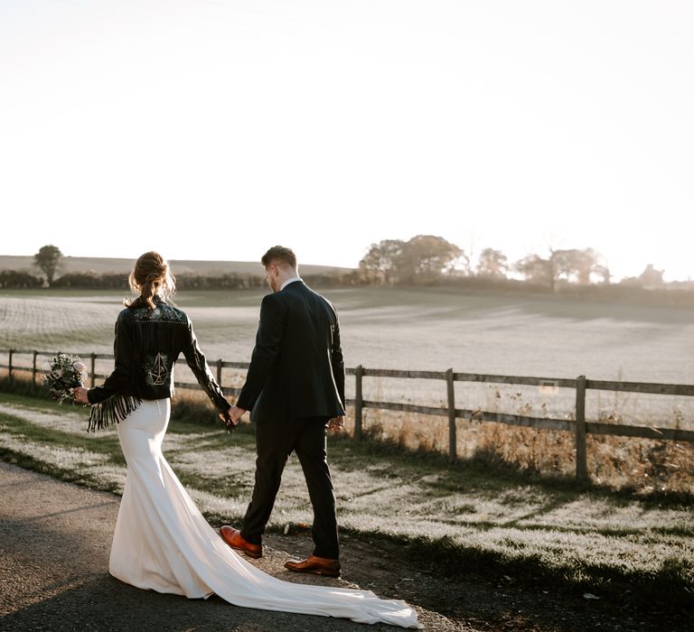 Bride and groom walking down a country lane with the bride in a fitted wedding dress and customised leather jacket with fringe sleeves and the groom in a navy suit