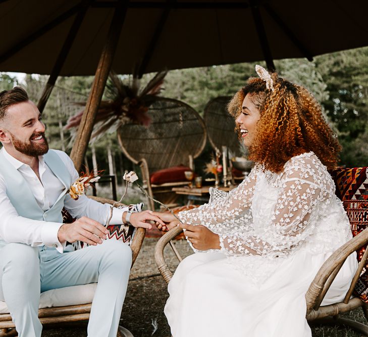 Bride & groom hold hands whilst sat on wooden chairs with Moroccan inspired cushions