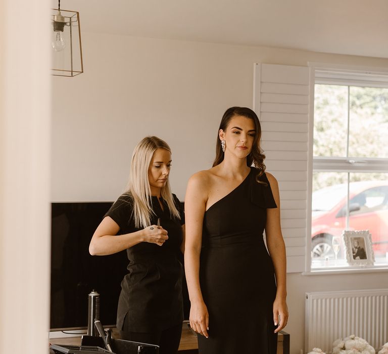 Bridesmaid in black off the shoulder bridesmaid dress with hair stylist in black tunic touching up hair before wedding