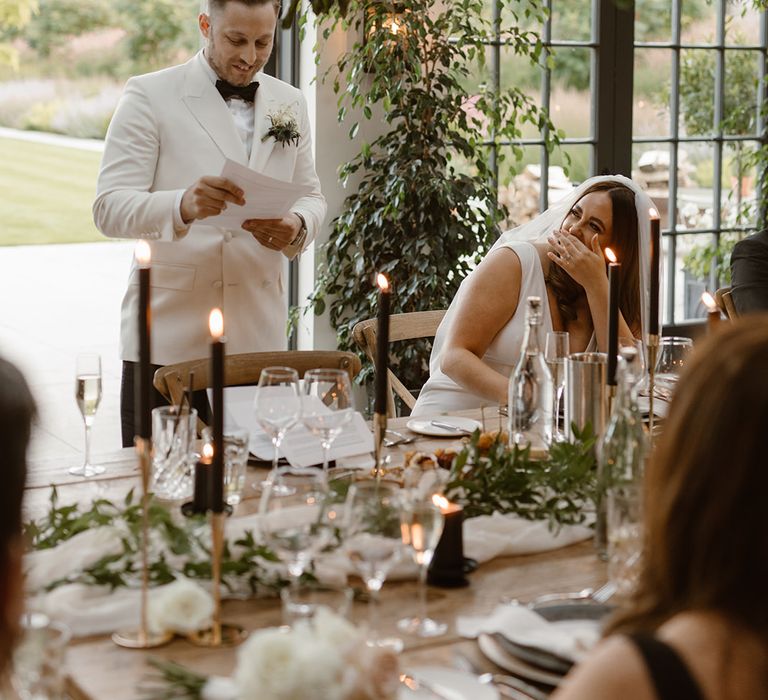 Groom in double breasted white tuxedo jacket giving speech in the Fig House at Middleton Lodge whilst bride in white Made With Love wedding dress looks up at him