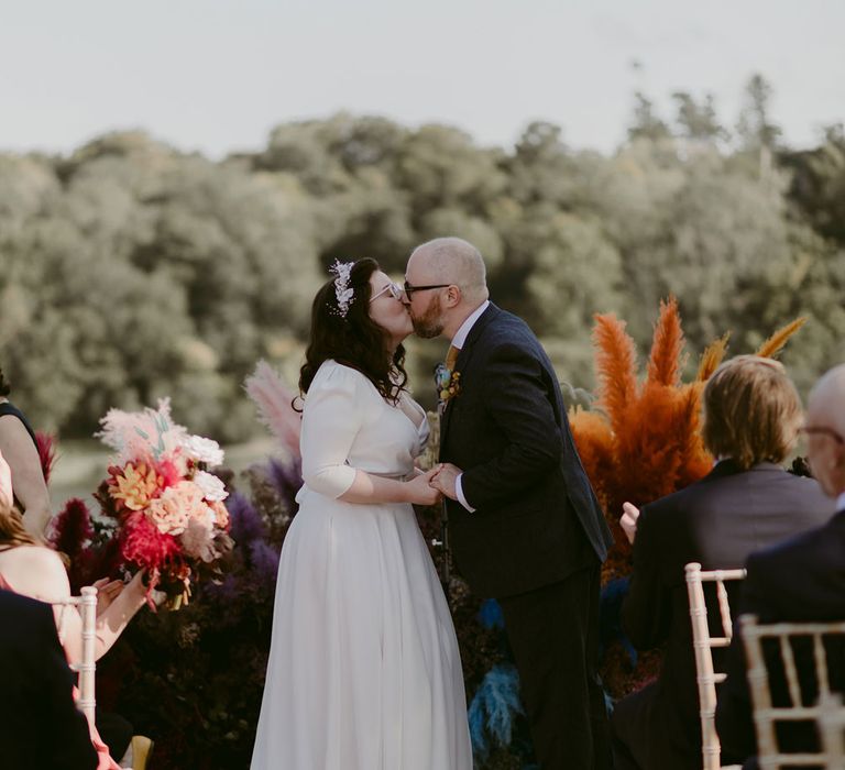 Bride in white Rime Arodaky wedding dress and bridal headband kisses groom in navy suit in front of multi-coloured pampas grass floral installation