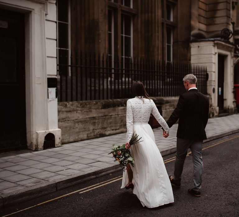 Bride in long sleeved lace top Self Portrait wedding dress carrying colourful bouquet holds hands with groom in brown woollen suit walking through streets of Bristol