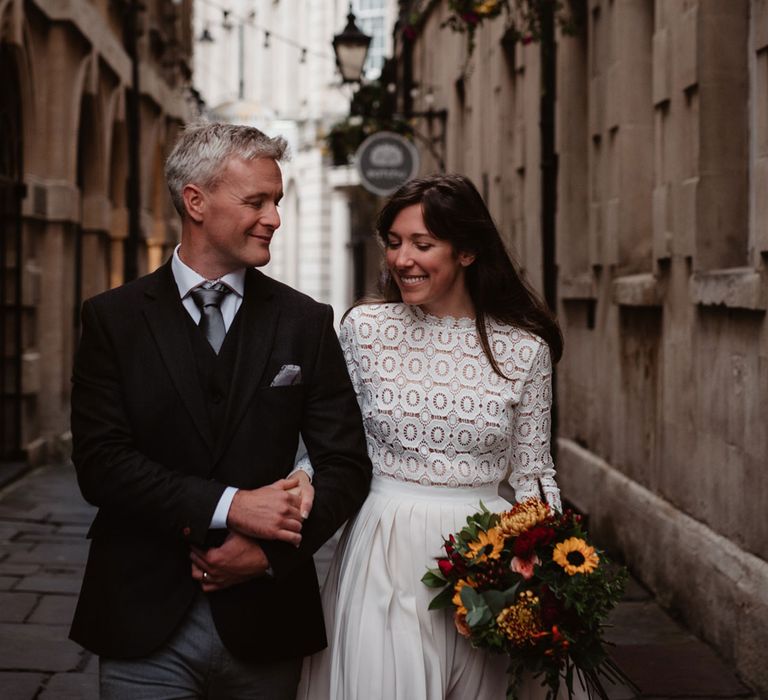 Bride in long sleeved lace top Self Portrait wedding dress carrying colourful bouquet holds hands with groom in brown woollen blazer walking through streets of Bristol