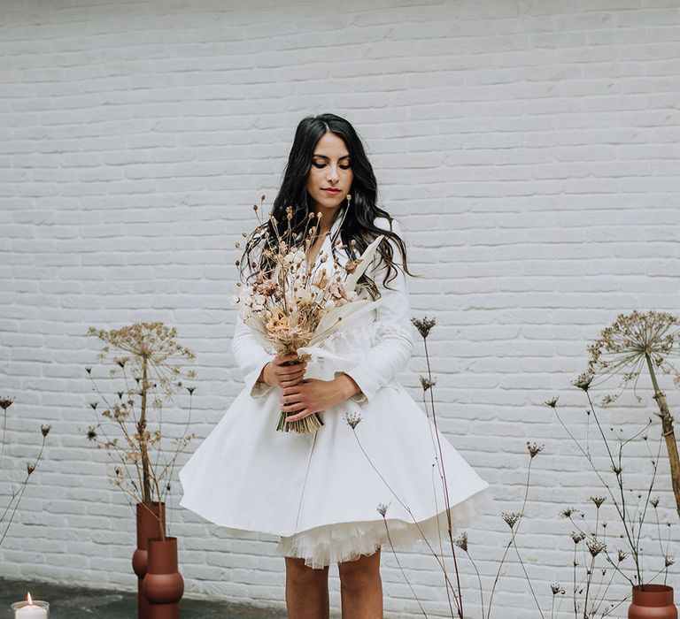 Bride standing in the candlelit aisle wearing a tutu blazer wedding dress and holding a minimal dried flower bouquet