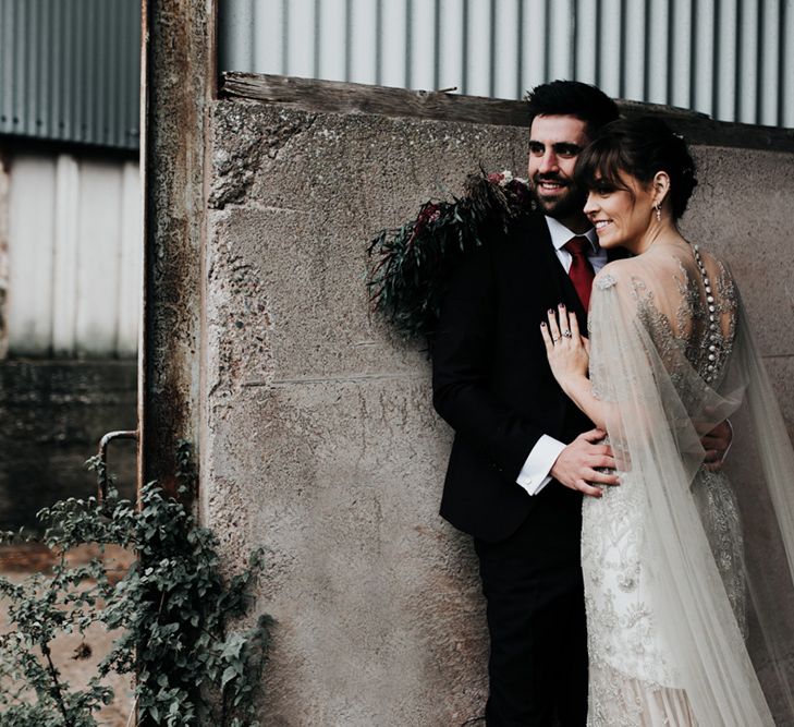 A bride and groom embrace. He leans against a wall. You can see the brides homemade wedding bouquet.