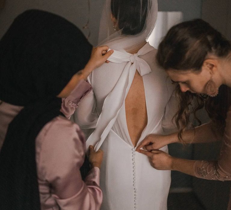 Wedding party helps bride with her gown as they do up buttons on her V backed dress