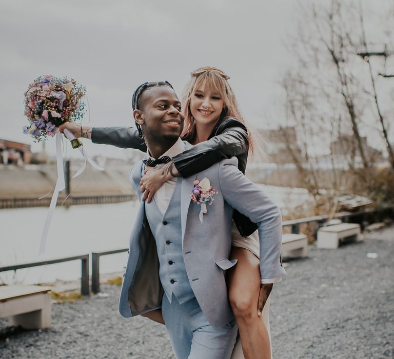 Groom in a pale blue three-piece wedding suit giving a piggy back to his bride in a leather jacket and boots 