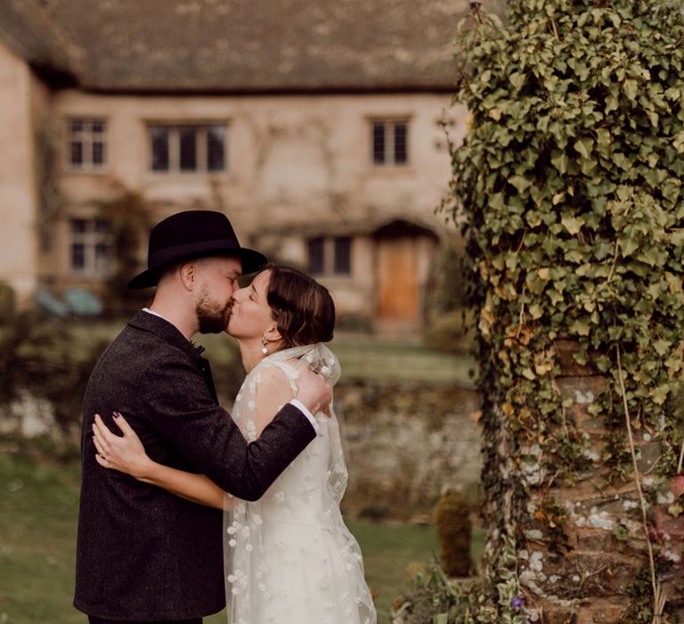 Bride in Charlie Brear wedding dress and daisy chapel length applique veil kisses groom in grey tweed suit and black fedora at garden party wedding at Hayne, Devon