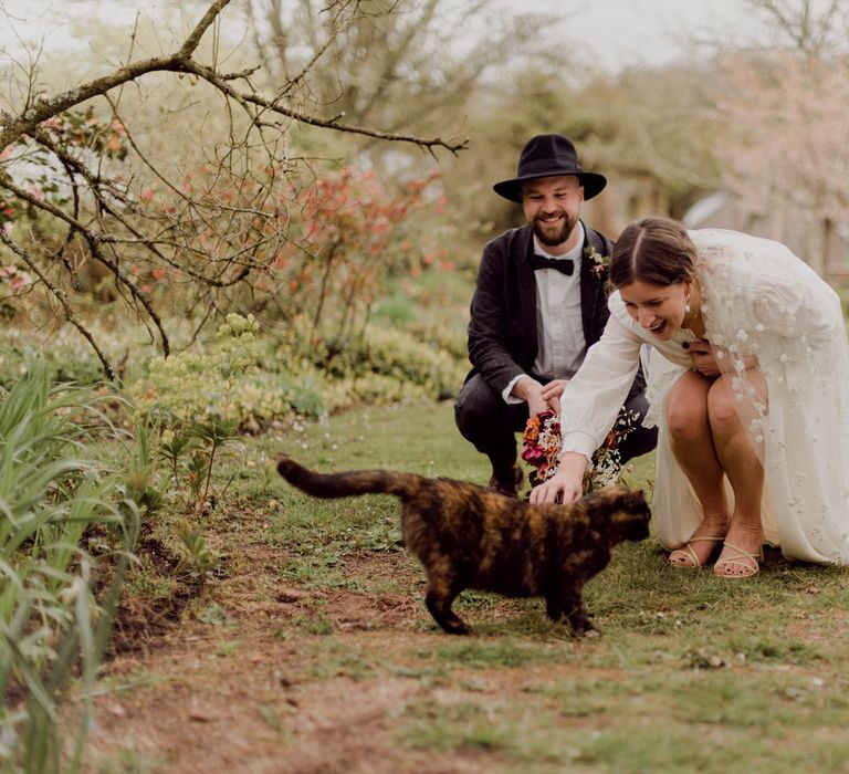 Smiling bride in white blouse, Charlie Brear wedding dress and applique veil holds multicoloured wedding bouquet strokes brown cat whilst kneeling alongside groom in brown tweed suit, bow tie and black fedora at garden party wedding in Devon