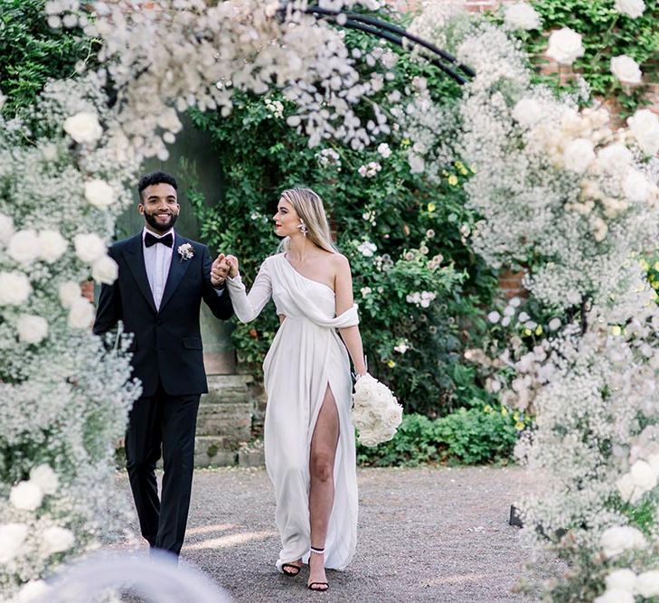 Groom in a black tuxedo and bow tie holding hands with this bride in a one shoulder wrap wedding dress in the gardens at Garthmyl Hall wedding venue 