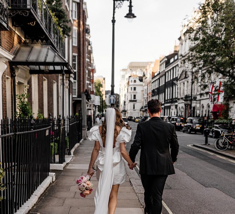 Bride in a short wedding dress with puff sleeves walking through Mayfair with her groom dresses in black with her long veil trailing behind them 