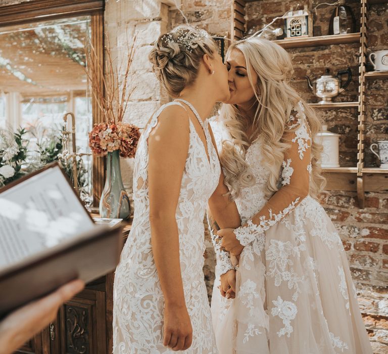 Two brides in white dresses with blonde hair kiss as a guest performs a reading at their wedding at Crab and Lobster Fairytale Wedding.