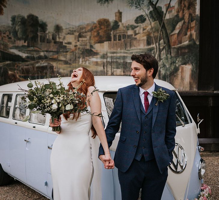 Groom in a three-piece navy suit and bride in a slip wedding dress holding a green and white bouquet in front of their VW camper van at the Larmer Tree wedding venue's outdoor theatre 