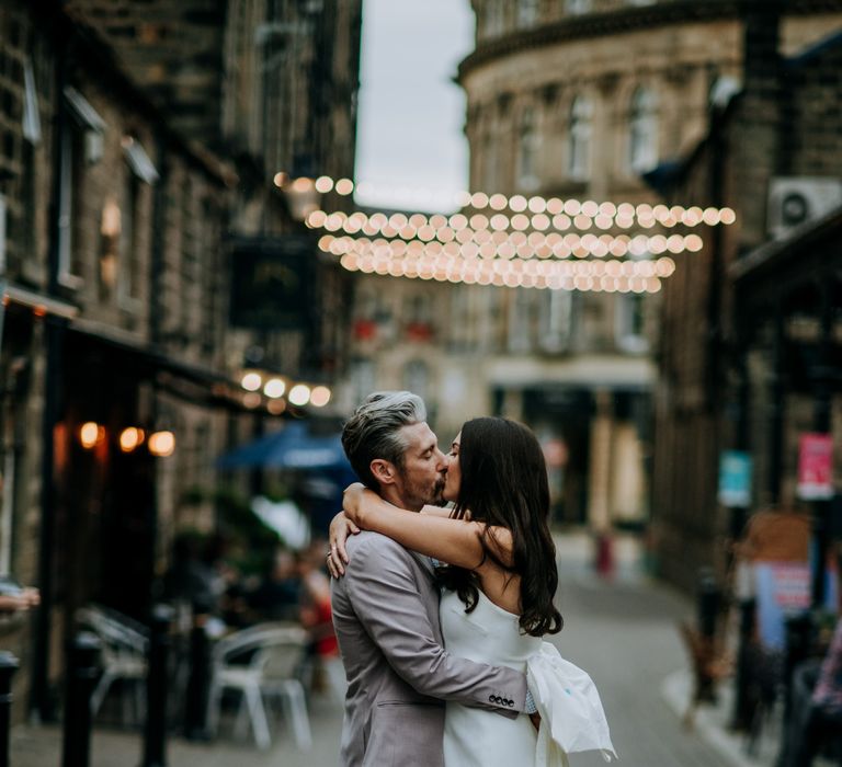 Bride in a strapless Rebecca Vallane Dress with giant bow on the back kissing her husband under a canopy of string lights