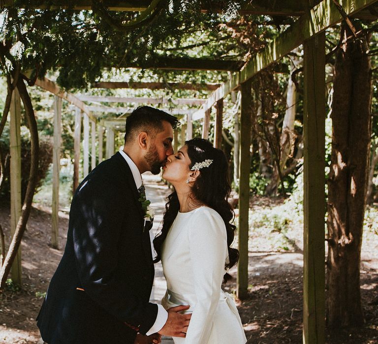 Groom in a navy blue suit kissing his bride in a long sleeve wedding dress under a pergola 