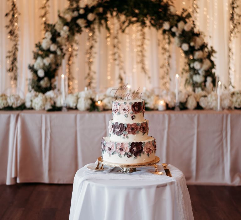 White frosted wedding cake sits in front of floral archway 