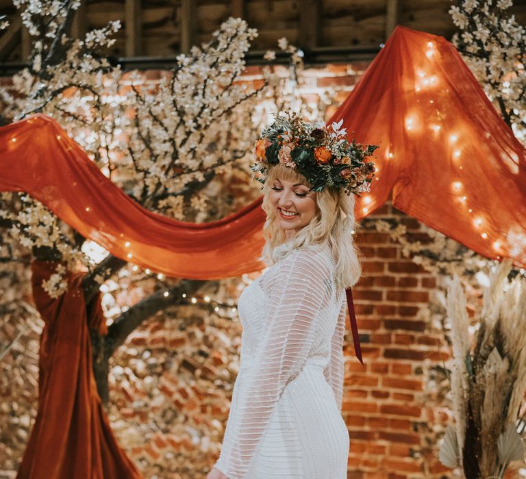 Bride wearing a long sleeve layered embroidered wedding dress with a huge flower crown in front of blossom trees draped in orange fabric