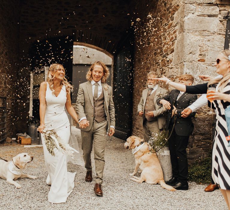 Bride in white V neck Made With Love wedding dress and veil holding white and green dried bridal bouquet walks through courtyard holding hands with groom in sage green suit and brown shoes as guests throw confetti after wedding ceremony at Anran Devon